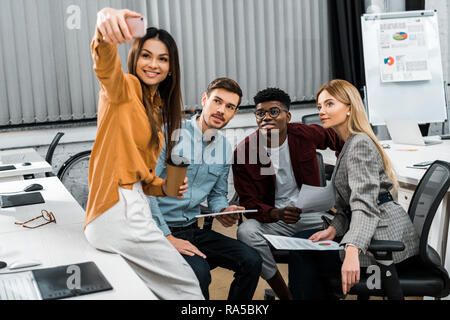 smiling multicultural businesspeople taking selfie on smartphone during work in office Stock Photo