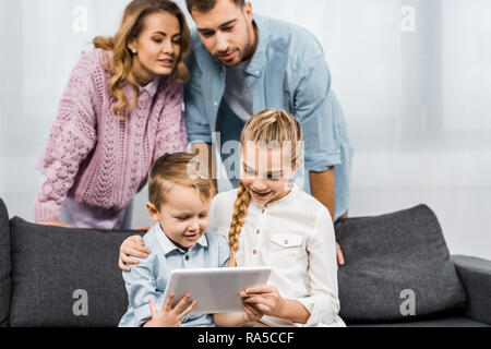 parents standing and looking at smiling siblings sitting on sofa and using digital tablet in living room Stock Photo