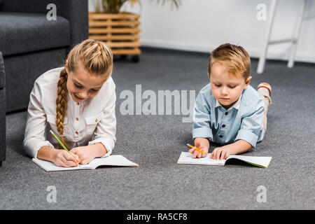 cute girl and boy lying on floor and writing in notebooks in living room Stock Photo
