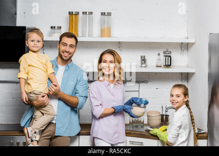 mother and daughter in rubber gloves washing dishes and looking at camera while father holding cute son in kitchen Stock Photo