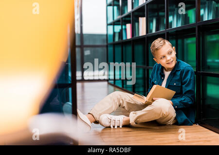 cute smiling boy reading book and looking away while sitting on floor in library Stock Photo