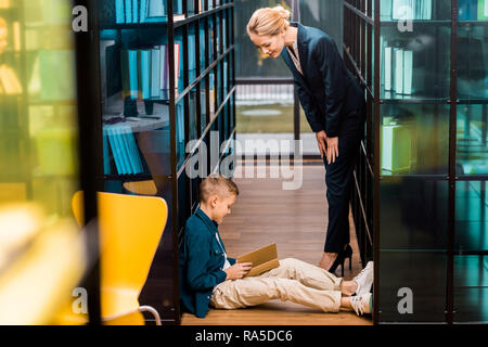 side view of young female librarian looking at boy reading book on floor in library