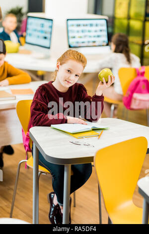 adorable schoolkid holding apple and looking at camera while sitting with classmates in library Stock Photo