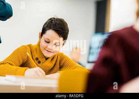 cute smiling schoolboy writing with pencil in classroom Stock Photo