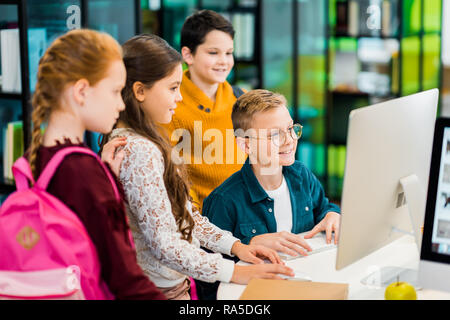 smiling schoolchildren using desktop computer together in library Stock Photo