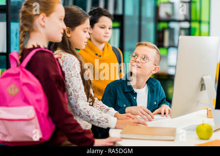 boy using desktop computer and looking at classmates in library Stock Photo