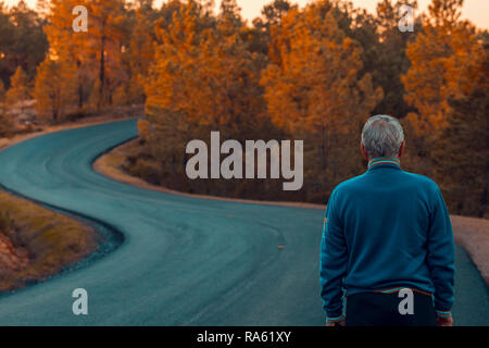 Active senior man stands alone on lonely road between mountains. Older man of back walking on lonely highway Stock Photo