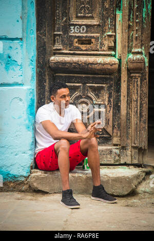 young Cuban man sitting on step in front of rustic door in a Havana Cuba street Stock Photo