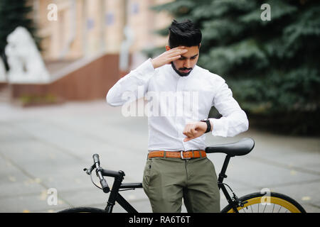 Smart indian businessman looking at his watch while late to meet by bicycle, copy space Stock Photo