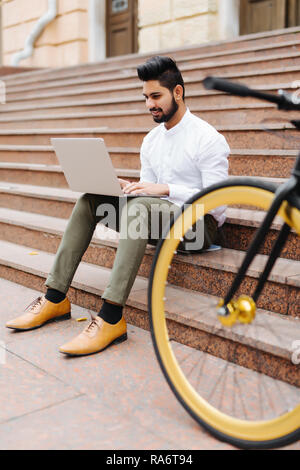 Young indian student working on laptop while sitting on stairs at the street Stock Photo