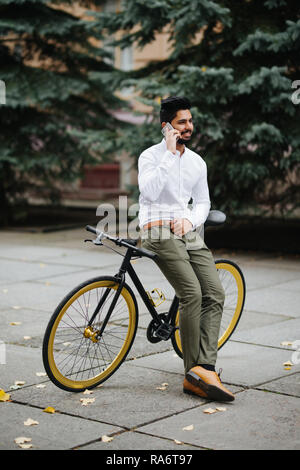 Handsome young indian man wearing suit talking on phone while walking with bicycle to work in the morning. Stock Photo