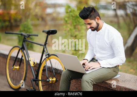 Confident young indian man working on laptop computer while sitting outdoors with bicycle Stock Photo