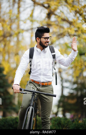 Casual handsome businessman going to work by bicycle. He is riding bike and waving Hello. Stock Photo