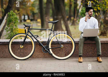 Happy young indian man in shirt talking on mobile phone while sitting with laptop computer outdoors with bicycle Stock Photo