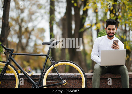 Happy young indian man using mobile phone while working on laptop computer sitting outdoors near bicycle Stock Photo