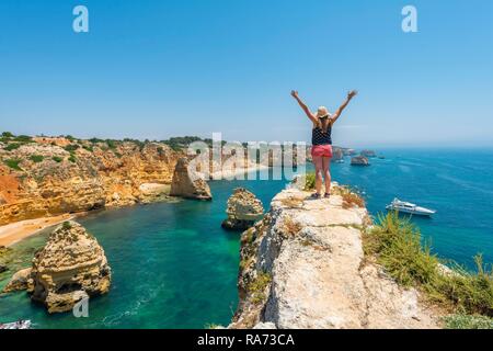 Young woman standing on rocks at steep coast, stretches arms into the air, view over turquoise sea, beach Praia da Marinha Stock Photo