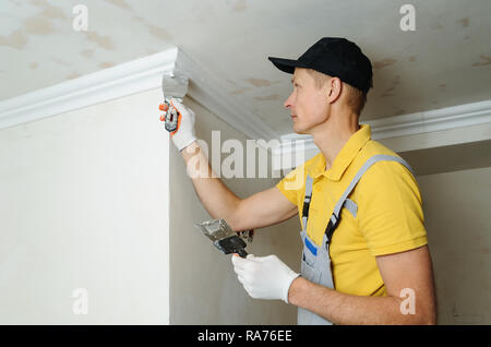 Installation of ceiling moldings. Worker puts glue on plastic molding for fixing it to the ceiling. Stock Photo
