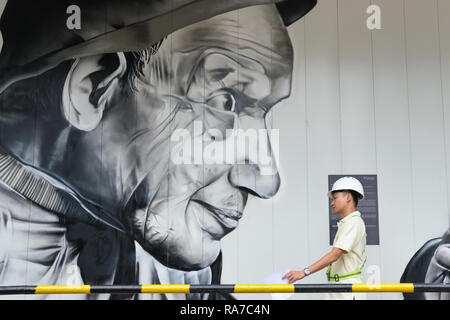 A construction engineer, seemingly observed by a painting of an oversized man, passing a make-shift wall shielding the construction site, Singapore Stock Photo