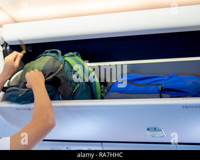 Passenger hand putting green backpack on locker number 10 in the economy class cabin in low cost airline airplane. Stock Photo