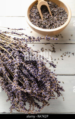 Bunch of dried lavender herbs resting on painted white wood planked table, with bowl of flower buds and woden scoop in soft focus background.. Stock Photo