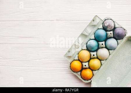 Stylish easter eggs in carton tray on white wooden background, top view. Modern colorful easter eggs painted with natural dye in different colors. Hap Stock Photo