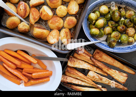 Roast potatoes, brussel sprouts, carrots and parsnips in baking trays and bowls with spoons on a slate background Stock Photo