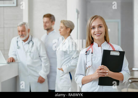 Young female doctor standing with tablet and looking at camera. Her colleagues standing behind her and discussing work searching solutions and help ill patients. Concept of health care. Stock Photo