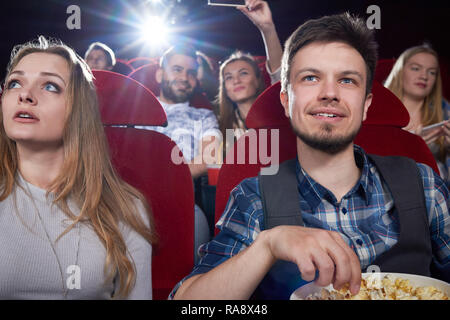 Front view of couple eating popcorn and looking at screen in cinema theater with red chairs. Blonde girl in gray and handsome man having romantic date and enjoying funny movie. Stock Photo