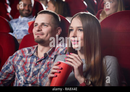 Couple of man in checked shirt and woman in gray sitting together in cinema, watching romantic movie. Handsome bearded man hugging beautiful girlfriend who sitting near and holding cup of drink. Stock Photo