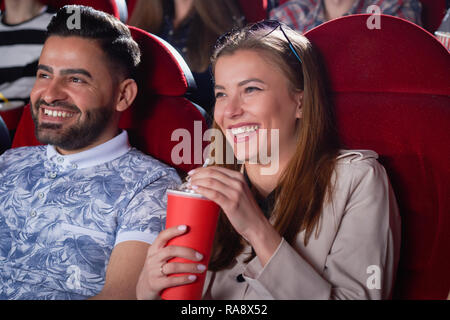 Positivity couple of blonde in gray and Arabian man in blue shirt drinking and smiling spending time in cinema. Students having fun, when looking at screen in modern cinema hall. Stock Photo