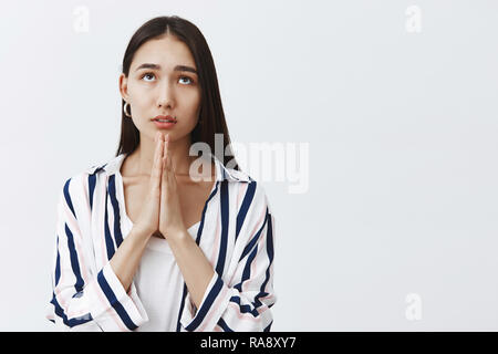 Girl praying god, asking for mercy and luck. Portrait of focused good-looking young woman in striped blouse, holding hands in pray, gazing up with dreamy expression, hoping, being believer Stock Photo