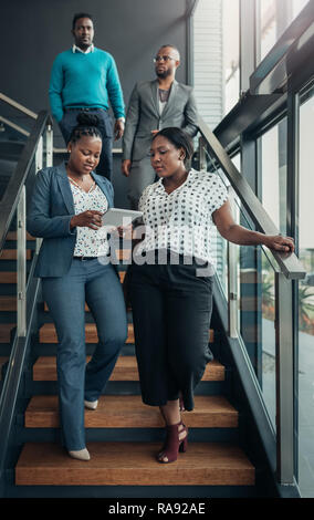 Team of african business people walking down a staircase after a meeting, the ladies in front are talking while using a tablet Stock Photo