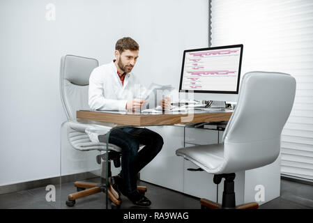 Handsome doctor reading some documents sitting in the luxury medical office with computer on the background Stock Photo