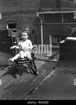 A toddler relaxes on a rooftop in a period wooden rocking chair,  ca. 1925. Stock Photo