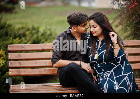 Indian couple posed outdoor, sitting on bench together and looking at ...