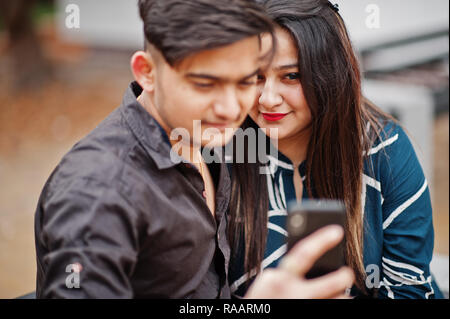 Indian couple posed outdoor, sitting on bench together and looking at mobile phone, making selfie. Stock Photo