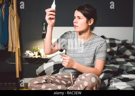 The theme is seasonal cold, runny nose flu virus infection. Young caucasian woman at home bedroom bed uses spray drops of medicine in her nose for sno Stock Photo