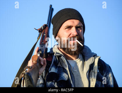 Guy bearded hunter spend leisure hunting and smoking. Hunting masculine hobby concept. Man brutal bearded guy gamekeeper blue sky background. Brutality and masculinity. Hunter with rifle gun close up. Stock Photo