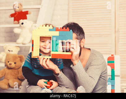 Dad and kid with toys on wooden background hold house wall made of plastic blocks. Father and son with happy faces play with colored toy bricks. Man and boy play together. Family and childhood concept Stock Photo