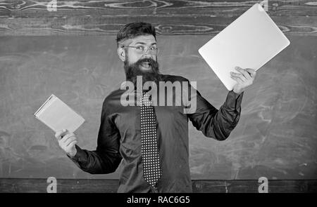 Teacher bearded hipster holds book and laptop. What would you prefer. Teacher choosing modern teaching approach. Paper book against laptop. Digital against paper. Choose right teaching method. Stock Photo