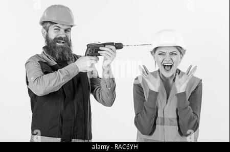 Husband annoying his wife. Man with happy face drills head of woman, white background. Builder, repairman makes hole in female head. Woman on scared face in helmet, hard hat. Annoying repair concept. Stock Photo