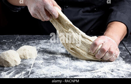 chef in black uniform kneads white wheat flour dough on a black wooden table Stock Photo