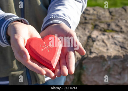 A boy holds and shows a wooden red heart in his hand, being in a park among trees on a sunny Valentine's Day and Mother's Day Stock Photo