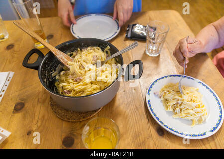 A pot full of pasta carbonara, two plates on a table. A young women is putting pasta on an empty plate. Stock Photo