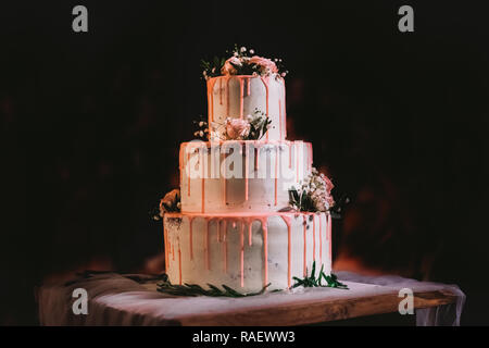 Beautiful elegant big three layers white wedding cake decorated with pink cream and fresh flowers. Cake standing on table isolated at black background Stock Photo