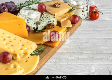 different types of cheeses on wooden cutting board. Camembert cheese, cheddar, hard cheese slices, soft cheese, walnuts, grapes, tomatoes and rosemary Stock Photo