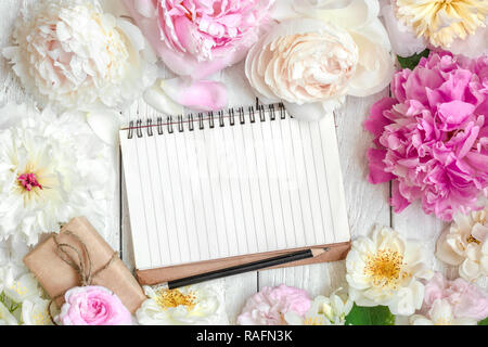 blank lined notebook in flower frame made of pink and white peony, roses and jasmine flowers and gift box on white wooden table. mock up. top view wit Stock Photo