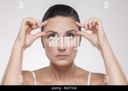 Portrait of pretty, charming, attractive middle aged woman with perfect face skin checking wrinkles on her forehead, holding fingers on brow, isolated Stock Photo