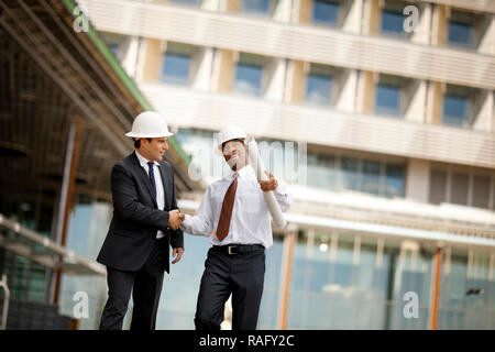 Two young engineers wearing suits and hardhats shaking hands after a discussion over building plans. Stock Photo