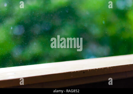 Summer rain raindrops falling down on wooden painted handle of balcony railings on blurred trees background Stock Photo